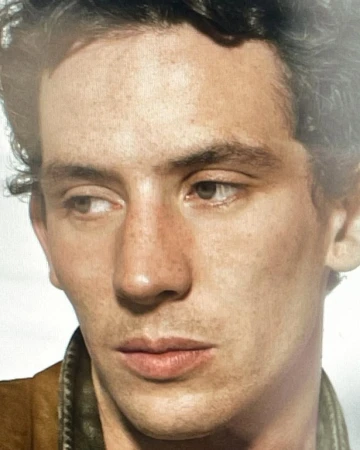 Close-up studio shot of a young adult male with freckles, dark curly hair, wearing a tan jacket and looking left.