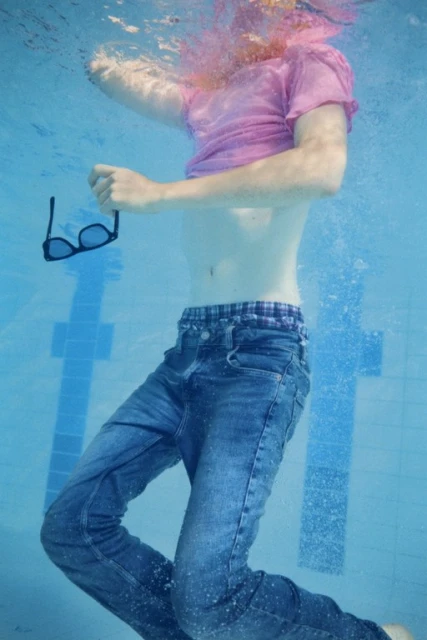 Underwater shot of a young adult in jeans and a pink t-shirt holding sunglasses in a blue swimming pool.