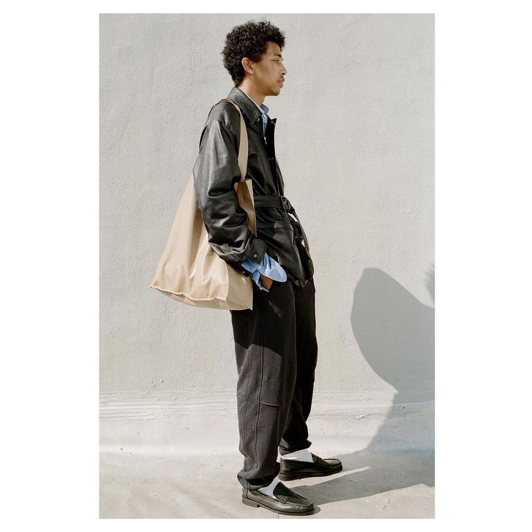 A profile view of a young man wearing a black leather jacket, dark trousers, and carrying a large beige tote bag.