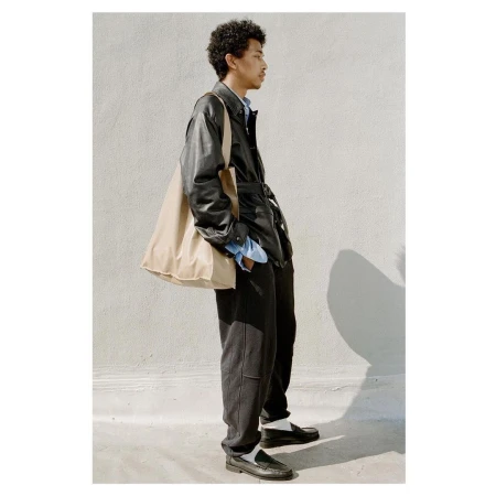 A profile view of a young man wearing a black leather jacket, dark trousers, and carrying a large beige tote bag.