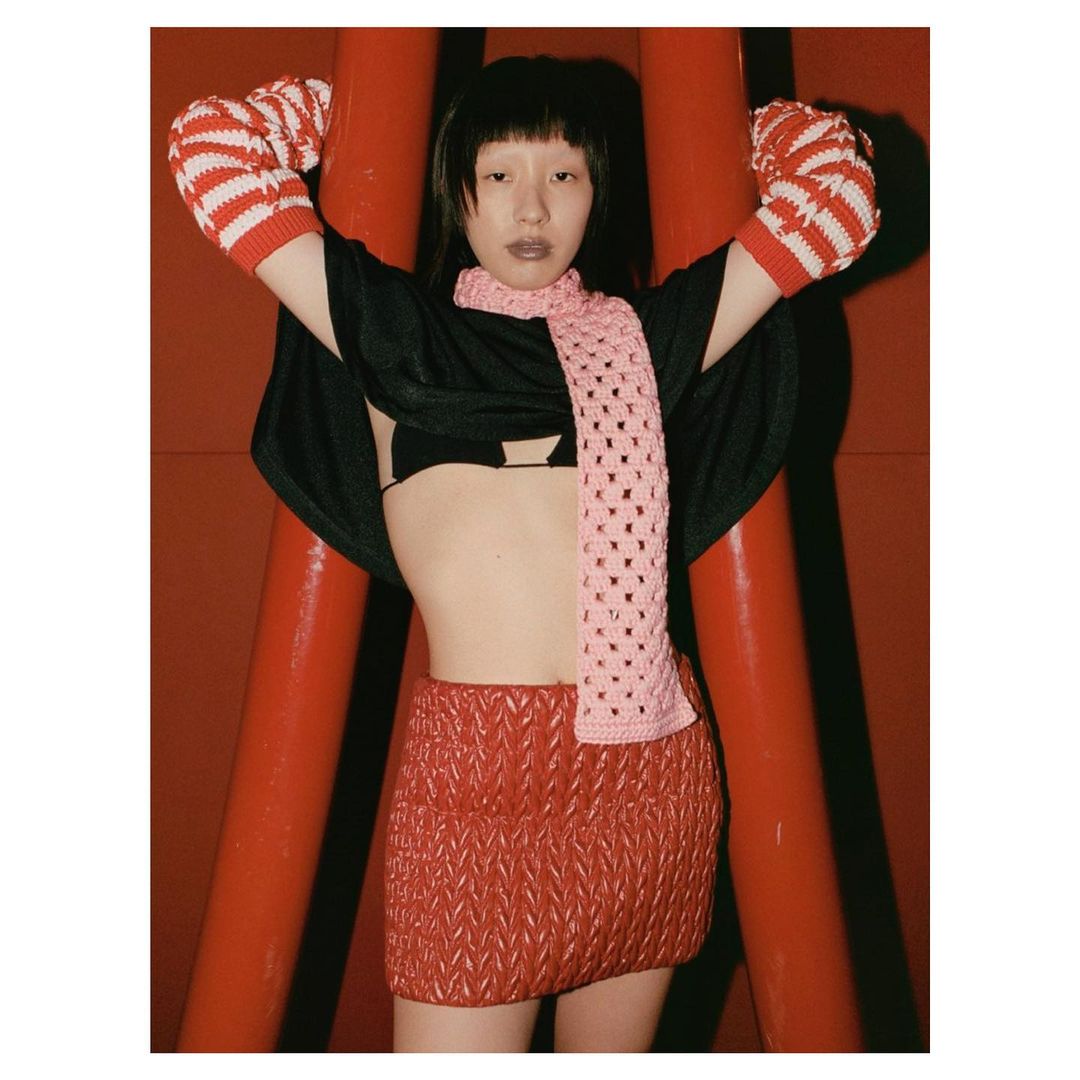 Woman in black top, red skirt and scarf posing against a red background with red and white striped arm warmers. Fashion editorial shot.