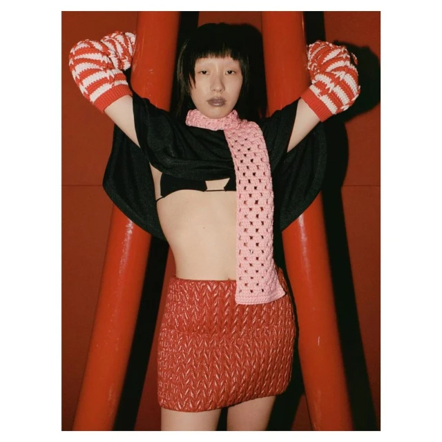 Woman in black top, red skirt and scarf posing against a red background with red and white striped arm warmers. Fashion editorial shot.