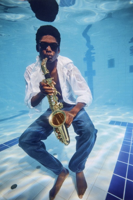 A man in jeans and a white shirt plays a saxophone underwater in a pool with blue and white tiles, creating a surreal musical moment.