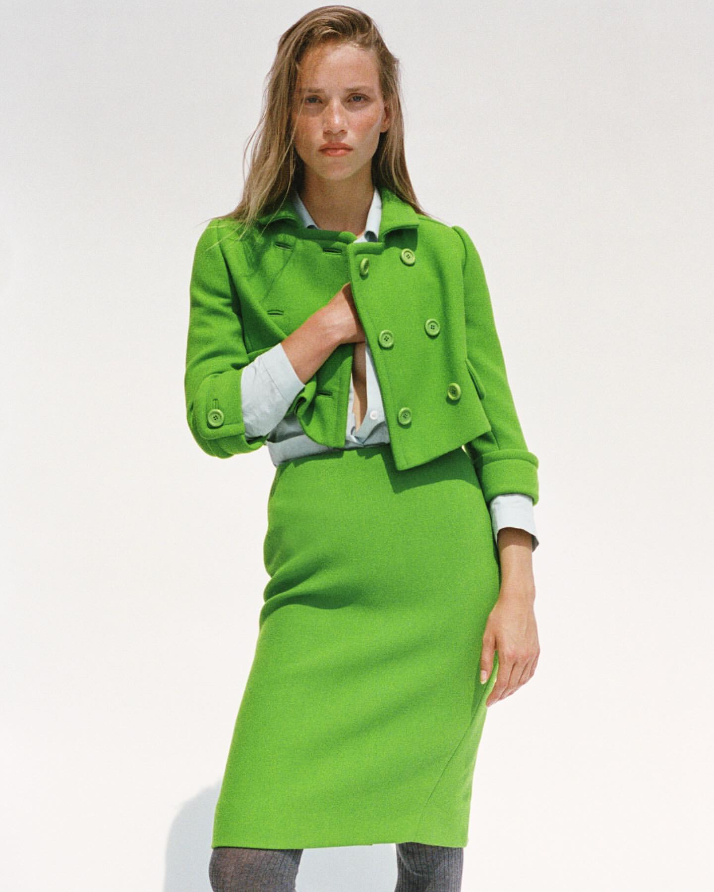 Adult woman poses in a bright green skirt suit in front of a white backdrop in a fashion photography style, showcasing the garment.