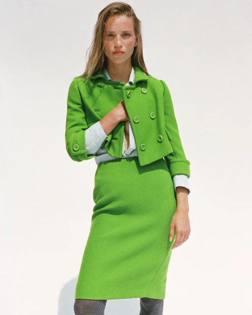 Adult woman poses in a bright green skirt suit in front of a white backdrop in a fashion photography style, showcasing the garment.