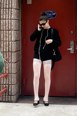 Woman in black sweater and pink thigh-high leg warmers talking on her phone outside a bright red door.