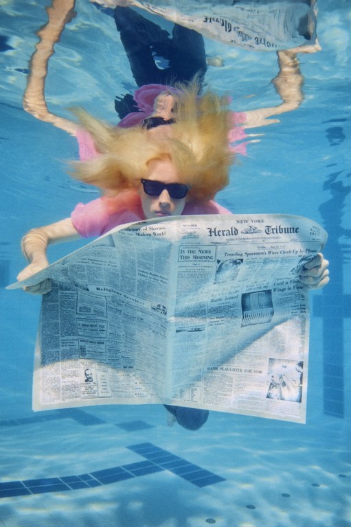 Underwater photo of a woman reading a newspaper in a swimming pool, wearing sunglasses and a pink shirt, with natural light.