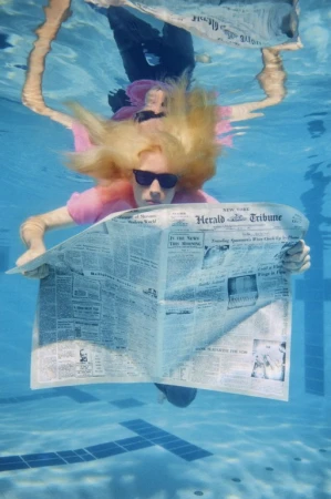 Underwater photo of a woman reading a newspaper in a swimming pool, wearing sunglasses and a pink shirt, with natural light.