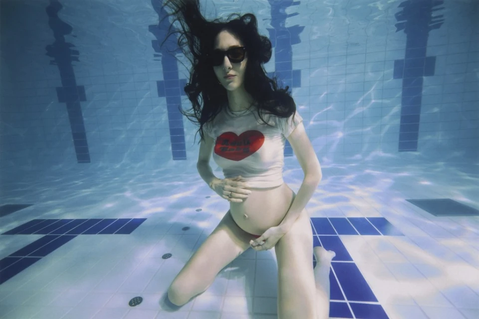 Underwater shot of a pregnant woman wearing sunglasses, a t-shirt with a red heart print, and red bottoms in a tiled swimming pool.
