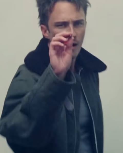 Close-up of an adult male wearing a dark green jacket with a black collar and a grey shirt, holding a small object in his fingers.