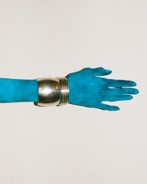 A turquoise arm extends horizontally across a white background, adorned with a thick, metallic silver cuff bracelet around the wrist.
