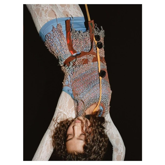 Upside-down view of a young adult female model with curly hair wearing a unique, handcrafted garment with red, white, and blue details.