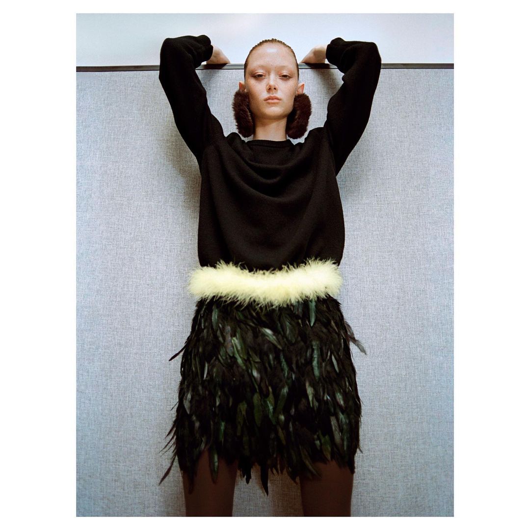 Fashion shot: A woman in a black sweater, feather skirt, and earmuffs leans against a gray wall, studio lighting.
