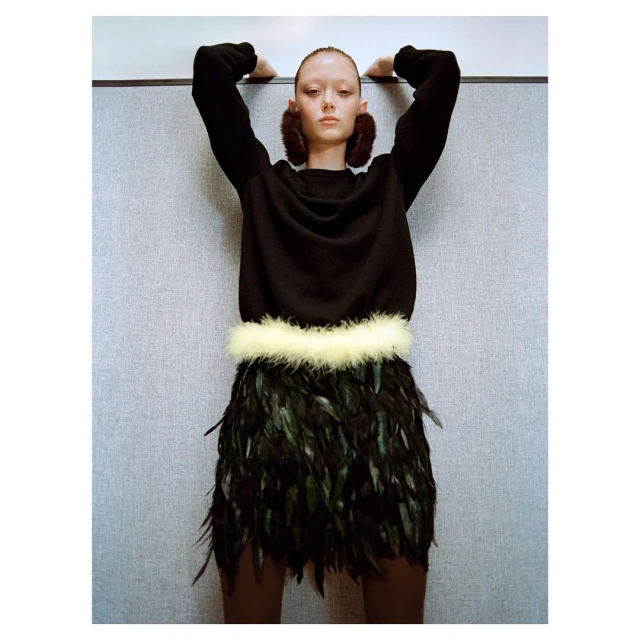 Fashion shot: A woman in a black sweater, feather skirt, and earmuffs leans against a gray wall, studio lighting.
