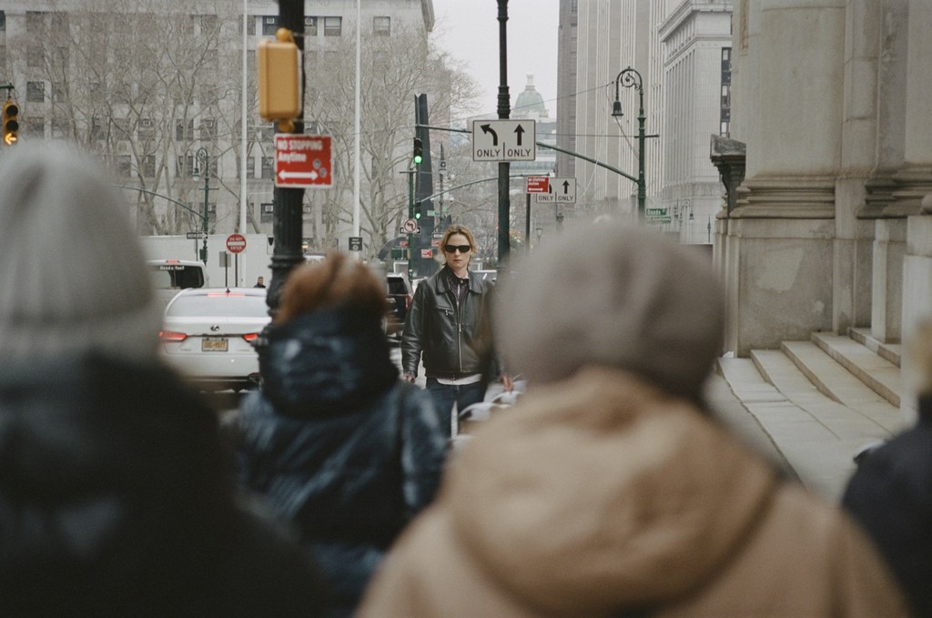 Adult female wearing sunglasses and leather jacket crossing the street in a city with buildings, traffic signals and other pedestrians.