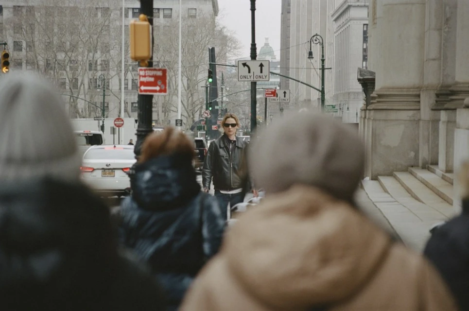Adult female wearing sunglasses and leather jacket crossing the street in a city with buildings, traffic signals and other pedestrians.