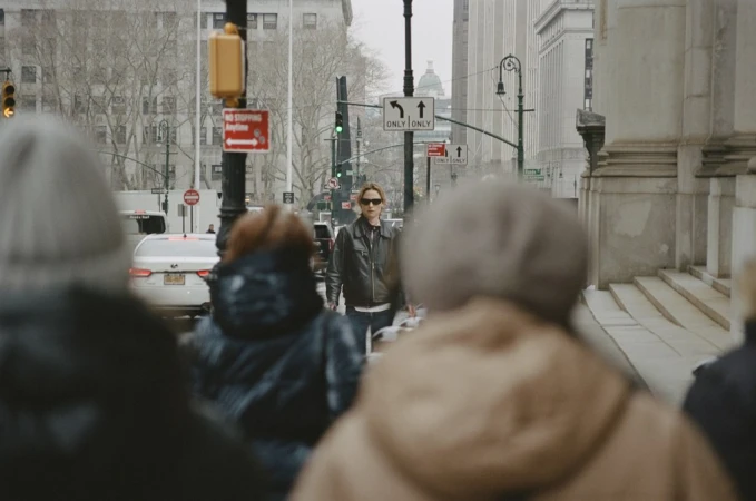Adult female wearing sunglasses and leather jacket crossing the street in a city with buildings, traffic signals and other pedestrians.