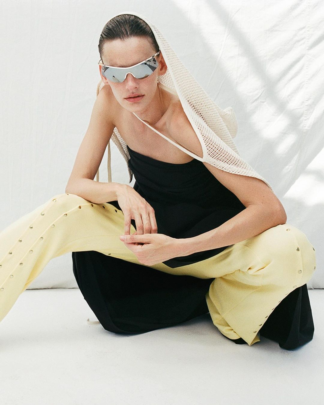 Adult woman with neutral expression poses in studio wearing black dress, cream pants with metal accents, silver sunglasses, and white netting.