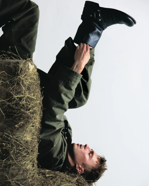 Adult male laying on a hay bale, wearing a green jacket, dark pants and a black boot while adjusting the boot's strap.