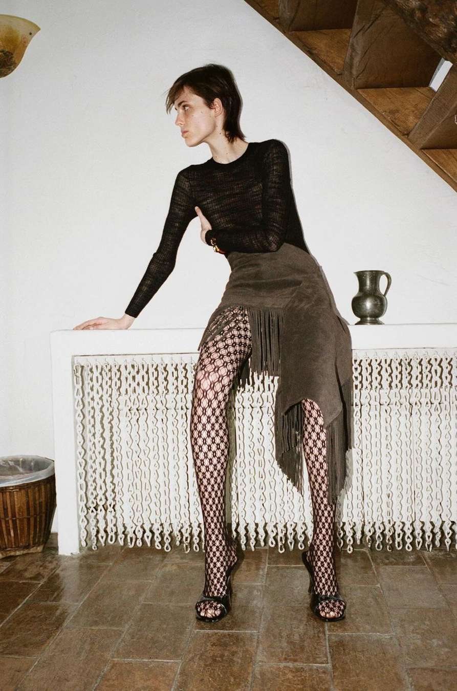 A woman poses indoors wearing a long-sleeved black shirt, an asymmetrical fringe skirt, and patterned tights next to a white fireplace.