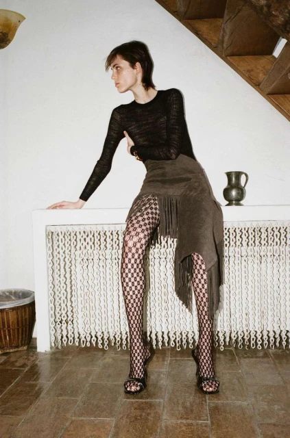 A woman poses indoors wearing a long-sleeved black shirt, an asymmetrical fringe skirt, and patterned tights next to a white fireplace.