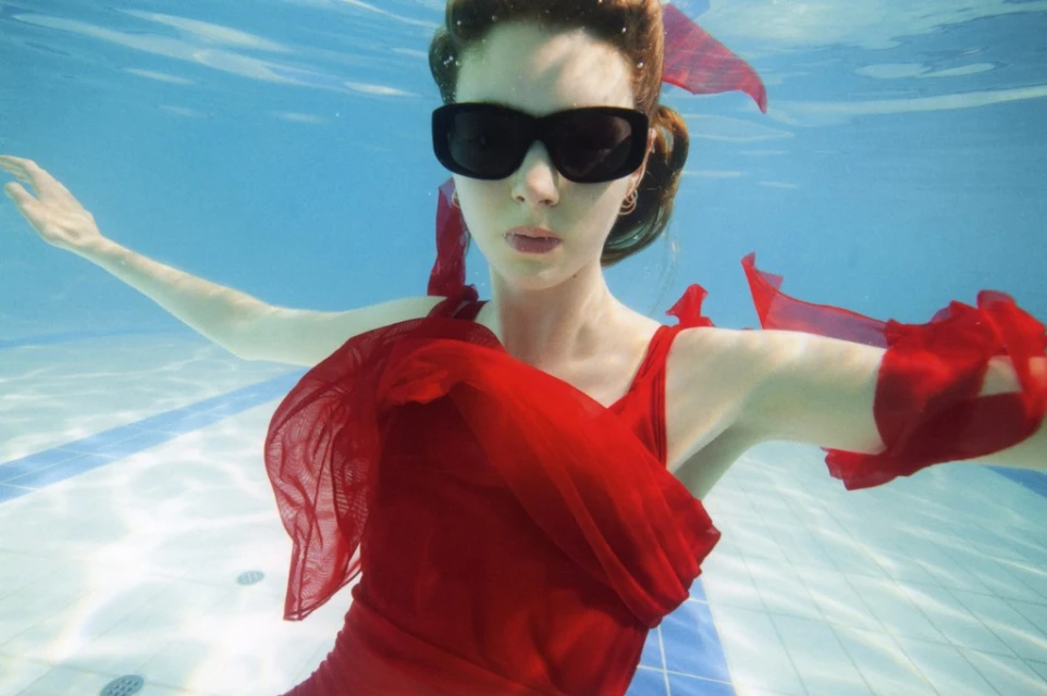 Woman in red dress and sunglasses underwater in a pool, red ribbons in hair, water ripples, and pool tiles visible, shot at eye-level angle.
