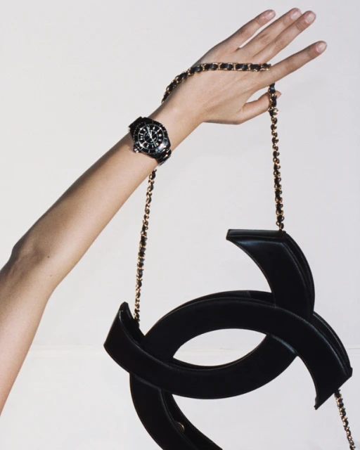 A black purse with a gold chain strap is being held up by a woman's hand. There is also a black watch on her wrist.