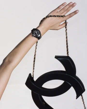 A black purse with a gold chain strap is being held up by a woman's hand. There is also a black watch on her wrist.