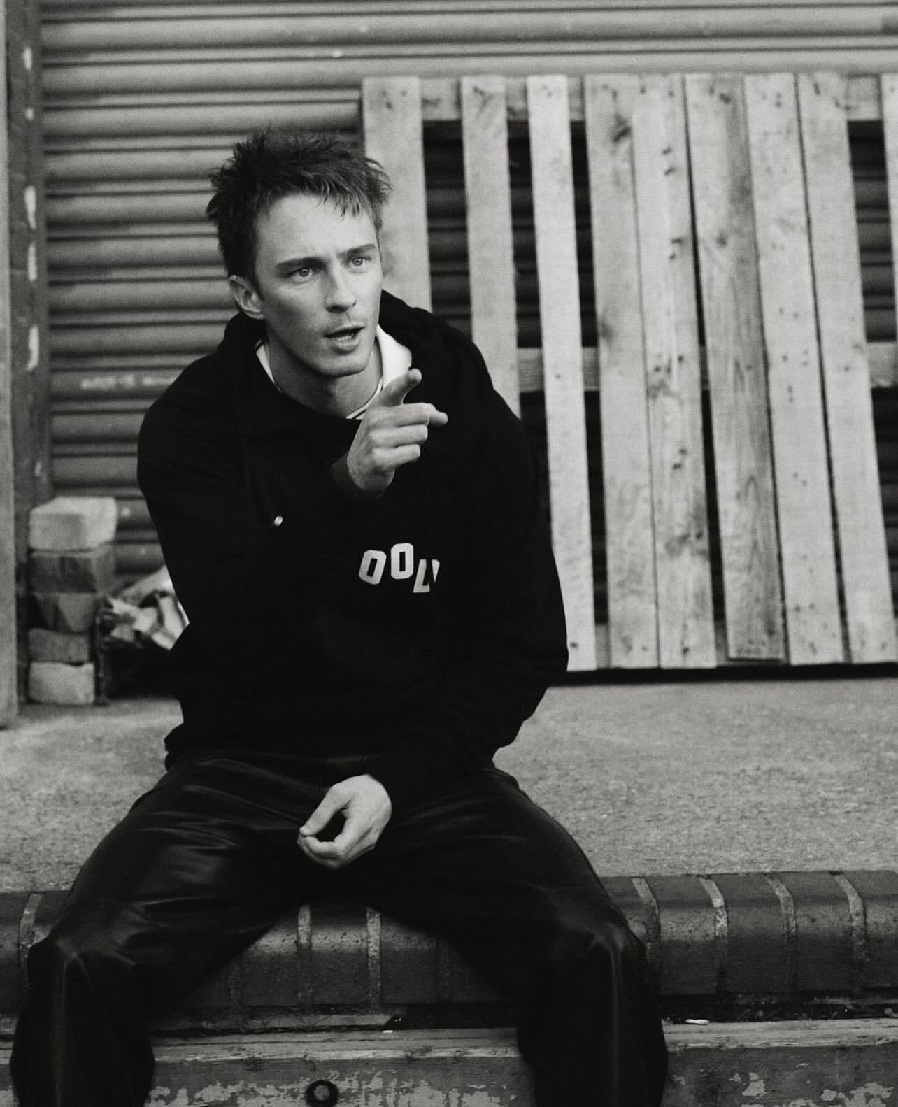 Monochrome portrait of a young adult male in a hooded sweatshirt, sitting on a brick ledge with a wooden pallet in the background.