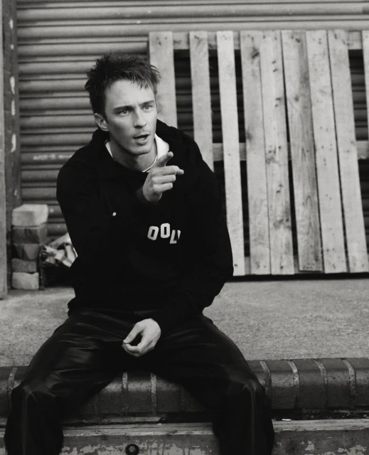 Monochrome portrait of a young adult male in a hooded sweatshirt, sitting on a brick ledge with a wooden pallet in the background.
