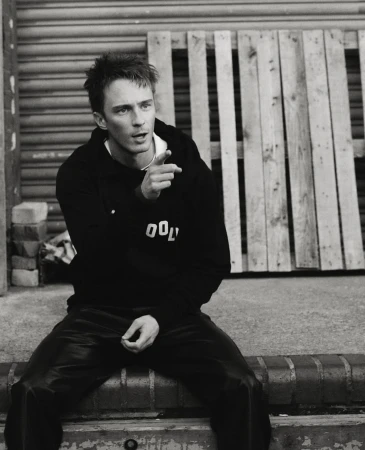 Monochrome portrait of a young adult male in a hooded sweatshirt, sitting on a brick ledge with a wooden pallet in the background.