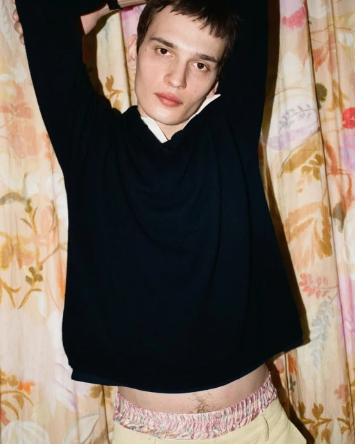 Young adult male in a dark sweater poses with arms up against a floral backdrop, casual indoor portrait, fashion lifestyle.