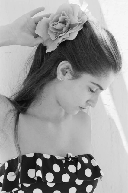 Black-and-white portrait of a young woman with long hair, accessorized with a large fabric flower, wearing a polka-dot top against a wall.