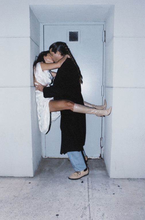 A romantic moment captured with a man holding a woman in his arms and kissing her in front of a pale blue door in an indoor setting.