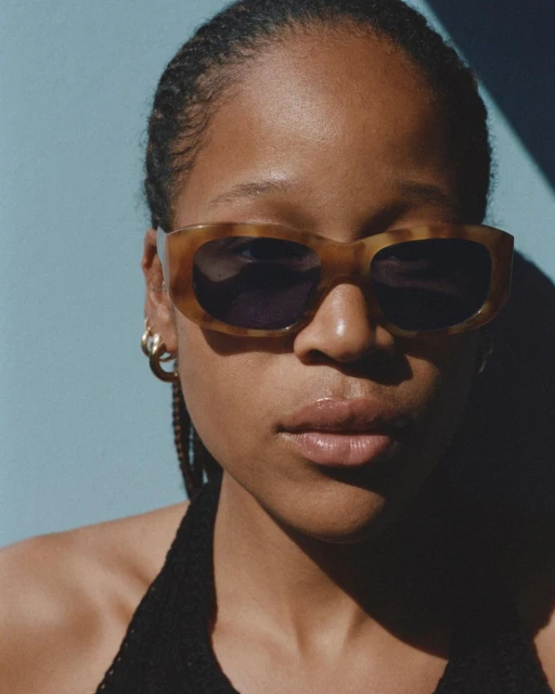 Close-up studio shot of a Black adult female model wearing sunglasses and a halter top against a blue background with strong shadow.