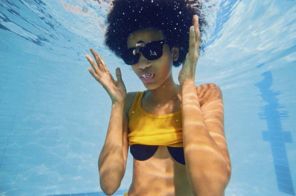 Underwater portrait of an African-American woman in sunglasses and a yellow swimsuit, with her hands raised, in a bright blue pool.