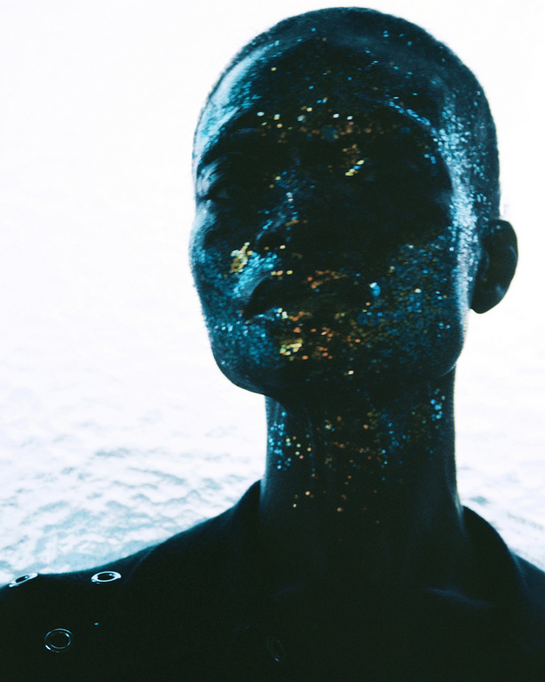 Close-up portrait of a bald adult female with glitter makeup, fair skin, wearing a black jacket and looking off to the side in a photo studio.