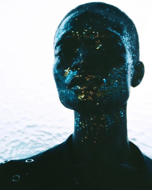 Close-up portrait of a bald adult female with glitter makeup, fair skin, wearing a black jacket and looking off to the side in a photo studio.