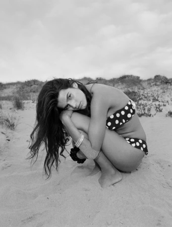 Woman in polka dot bikini crouching on sandy beach with her head resting on her knees