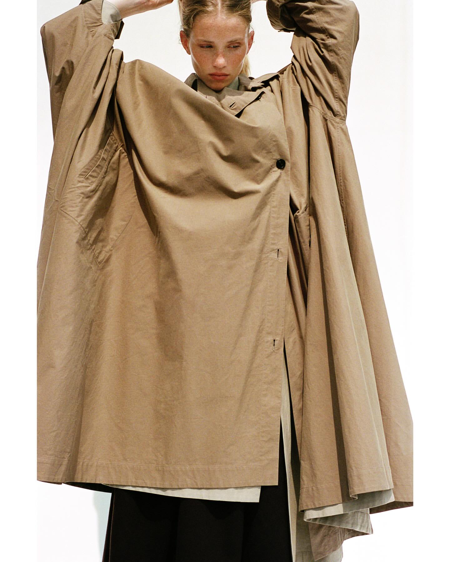 Fashion editorial shot of a fair-skinned adult woman wearing an oversized khaki coat, standing against a white backdrop in a photo studio.