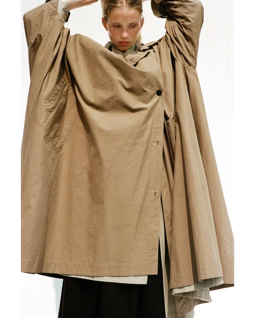 Fashion editorial shot of a fair-skinned adult woman wearing an oversized khaki coat, standing against a white backdrop in a photo studio.