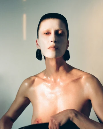Close-up portrait of a model with sleek dark hair, pale skin, and prominent dark beaded earrings against a soft neutral background.