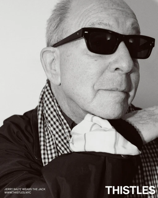 Portrait of Jerry Saltz wearing sunglasses and a checkered scarf. The image is black and white, with the brand THISTLES visible.