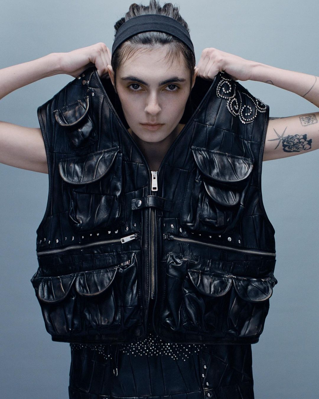 Woman poses indoors wearing a black leather studded vest with pockets. The model has a headband and tattoos on her arms.