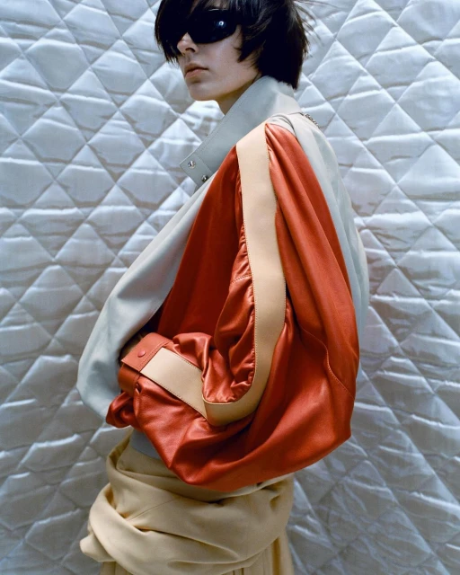 Fashion shot of a person with dark hair wearing sunglasses and a jacket with red sleeves posing against a quilted background indoors.