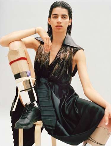 Fashion portrait of a model with a leg brace, sitting on a wooden stool against a white background, wearing a black dress and black sneakers.