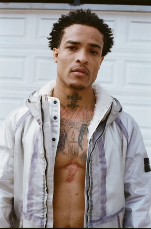 Portrait of a tatted man in a white zip-up jacket with an open chest posing in front of a white garage door outdoors.