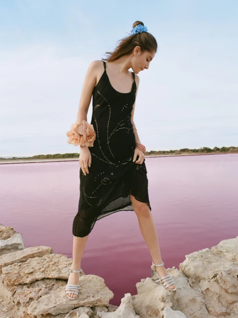 Young woman in a black dress and sandals standing on rocks at a pink lake under a bright sky, fashion editorial photography.