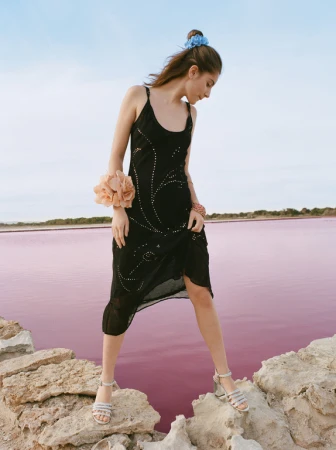 Young woman in a black dress and sandals standing on rocks at a pink lake under a bright sky, fashion editorial photography.
