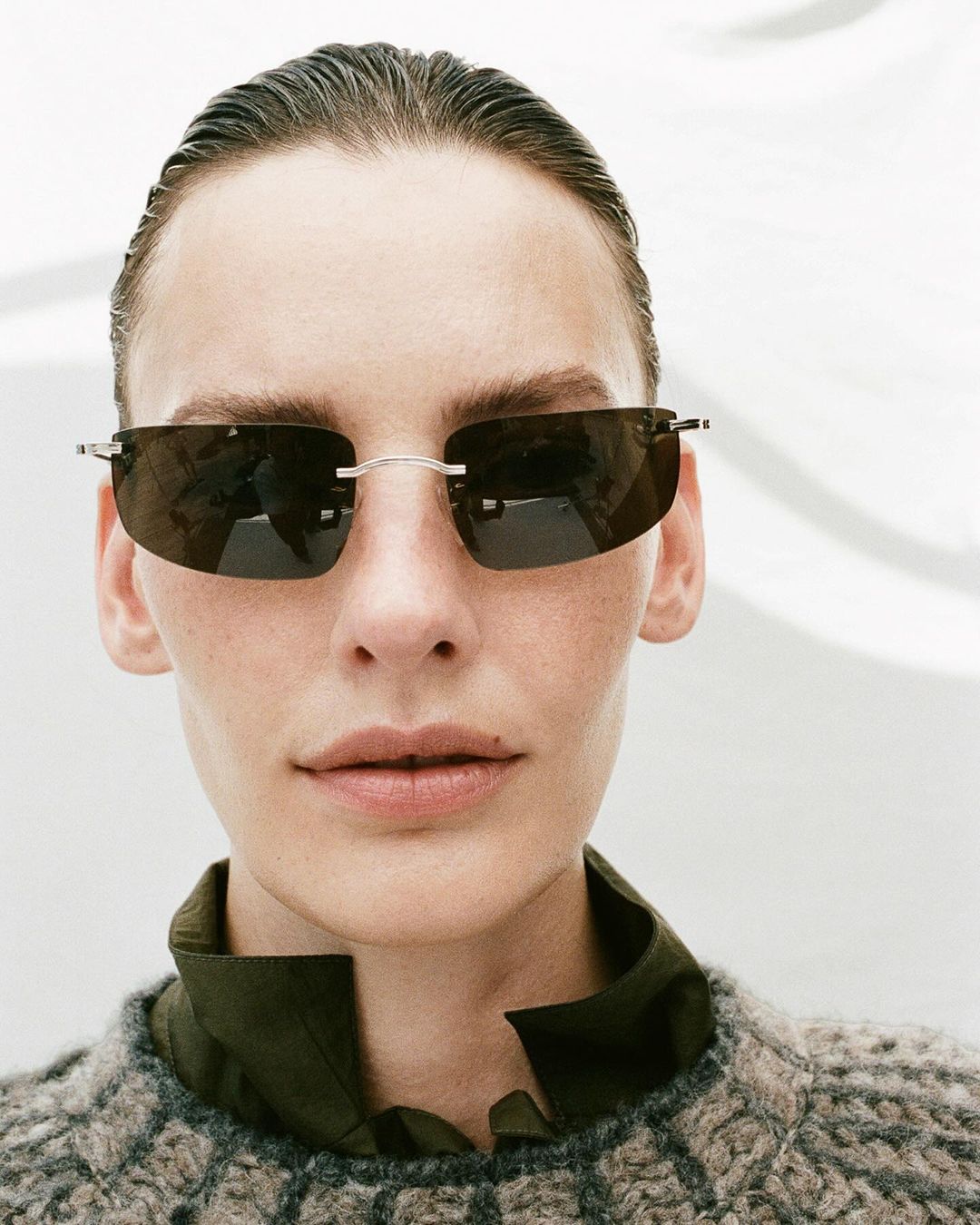 Close-up of adult woman wearing sunglasses, gray knit sweater, and dark green collared shirt against a blurry white background.